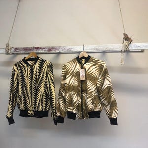 Puede incluir: Dos chaquetas bomber estampadas colgadas de un estante de madera. La chaqueta de la izquierda tiene un estampado de hojas en blanco y negro. La chaqueta de la derecha tiene un estampado de hojas en marr&oacute;n y blanco.