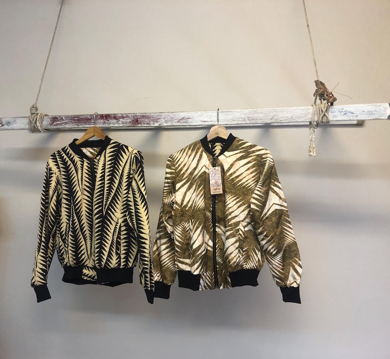 Puede incluir: Dos chaquetas bomber estampadas colgadas de un estante de madera. La chaqueta de la izquierda tiene un estampado de hojas en blanco y negro. La chaqueta de la derecha tiene un estampado de hojas en marr&oacute;n y blanco.