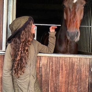 Könnte beinhalten: Eine Frau in einer grünen Kapuzenjacke mit einem braunen Pferd in einem Stall hinter ihr.