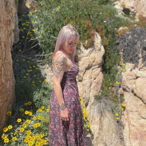 May include: A woman with blonde hair wearing a floral print maxi dress. She is standing on a rocky cliff with yellow flowers in the foreground.