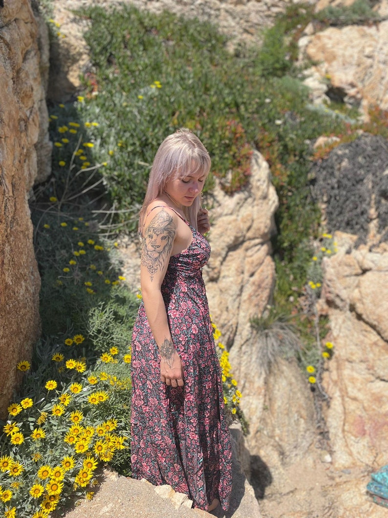 May include: A woman with blonde hair wearing a floral print maxi dress. She is standing on a rocky cliff with yellow flowers in the foreground.