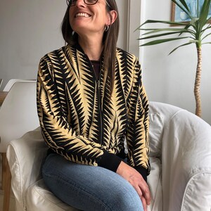 Puede incluir: Una mujer con una chaqueta bomber de color negro y amarillo con un patr&oacute;n de dise&ntilde;o. Est&aacute; sentada en una silla blanca y lleva pantalones vaqueros azules.