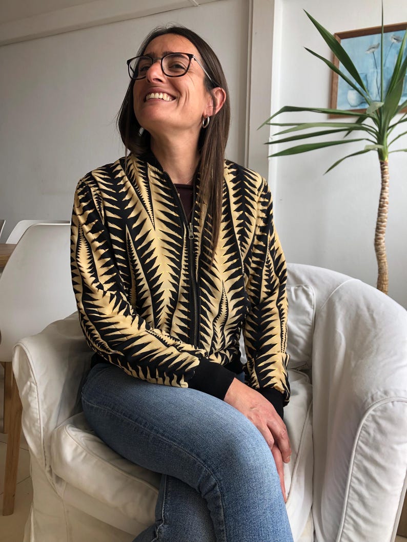 Puede incluir: Una mujer con una chaqueta bomber de color negro y amarillo con un patr&oacute;n de dise&ntilde;o. Est&aacute; sentada en una silla blanca y lleva pantalones vaqueros azules.
