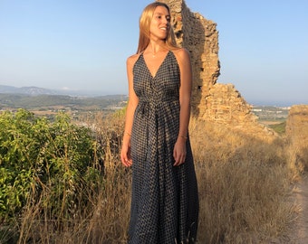 May include: A woman wearing a long, black and gold patterned dress with a halter neckline. She is standing in a field of tall grass with a stone wall in the background.