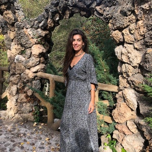 May include: A woman wearing a long, black and white floral print dress with a long sleeve and a V-neckline. She is standing in front of a stone archway.
