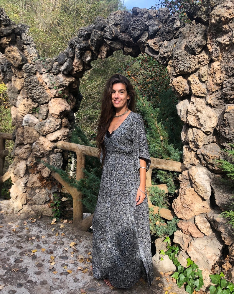 May include: A woman wearing a long, black and white floral print dress with a long sleeve and a V-neckline. She is standing in front of a stone archway.