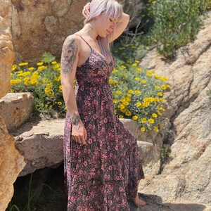 May include: A woman with pink hair wearing a black and pink floral maxi dress. The dress has a V-neckline and a tiered skirt. She is standing in front of a rocky background with yellow flowers.