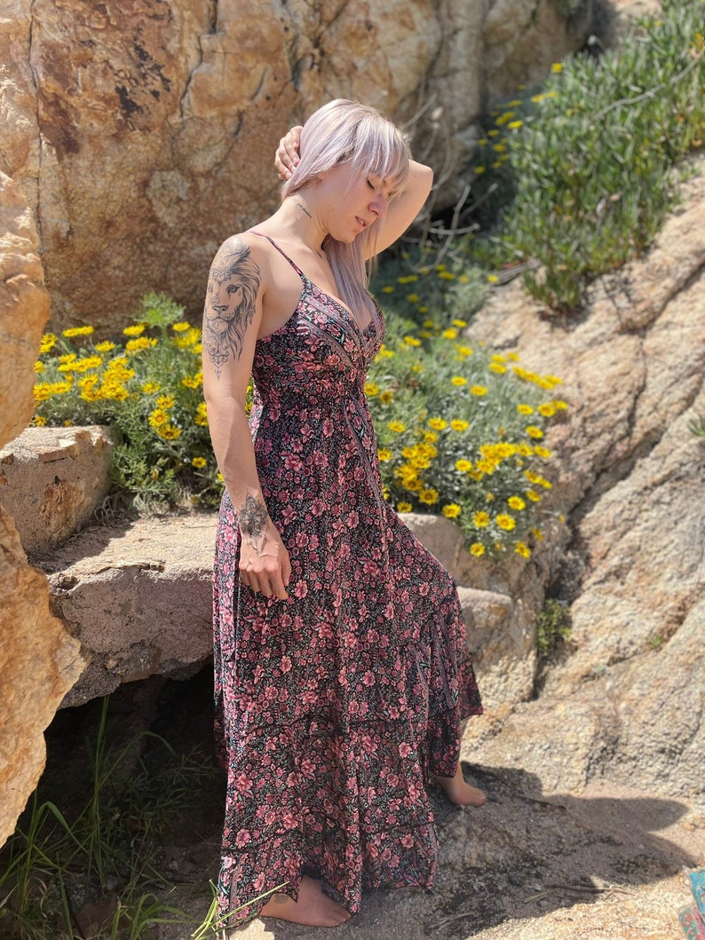 May include: A woman with pink hair wearing a black and pink floral maxi dress. The dress has a V-neckline and a tiered skirt. She is standing in front of a rocky background with yellow flowers.