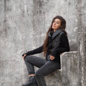K&ouml;nnte beinhalten: Eine Frau in einer schwarzen Jacke mit Muster sitzt auf einer Betontreppe. Sie tr&auml;gt eine schwarze Jeans und schwarze Stiefel.