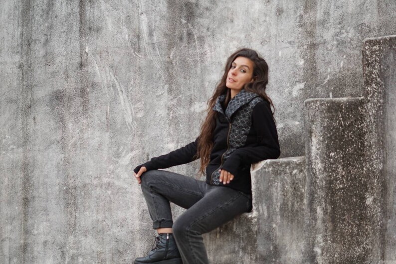K&ouml;nnte beinhalten: Eine Frau in einer schwarzen Jacke mit Muster sitzt auf einer Betontreppe. Sie tr&auml;gt eine schwarze Jeans und schwarze Stiefel.
