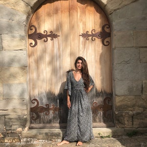 May include: A woman wearing a long, gray and white patterned dress with a V-neck and bell sleeves stands in front of a wooden door with ornate iron hinges.