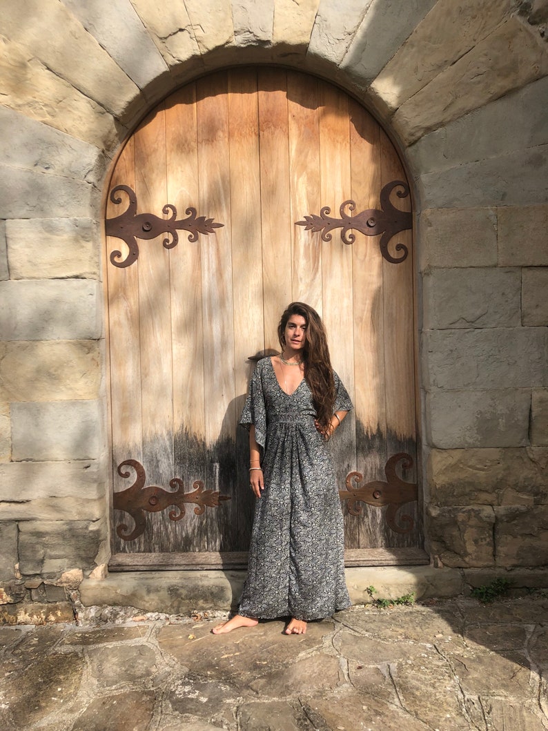 May include: A woman wearing a long, gray and white patterned dress with a V-neck and bell sleeves stands in front of a wooden door with ornate iron hinges.