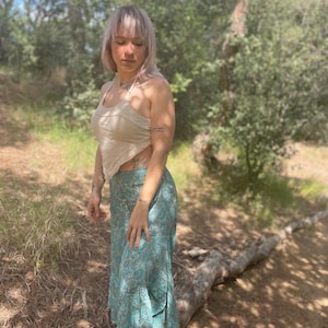 May include: A woman wearing a white halter top and a long, flowing teal skirt with a floral pattern. She is standing in a wooded area with a log behind her.