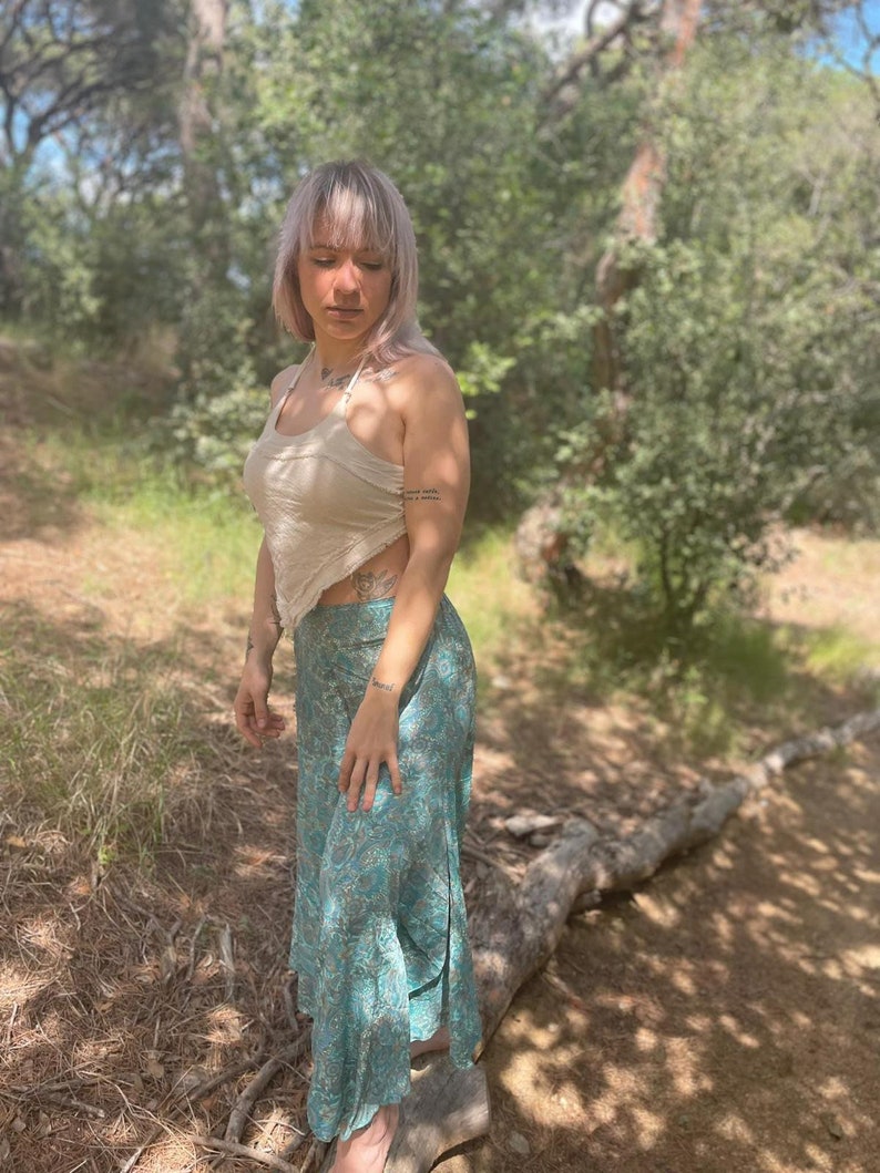 May include: A woman wearing a white halter top and a long, flowing teal skirt with a floral pattern. She is standing in a wooded area with a log behind her.
