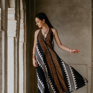 May include: A woman wearing a black and white patterned maxi dress with orange accents. The dress has a halter neckline and a flowing skirt. She is standing in a doorway with a concrete wall behind her.