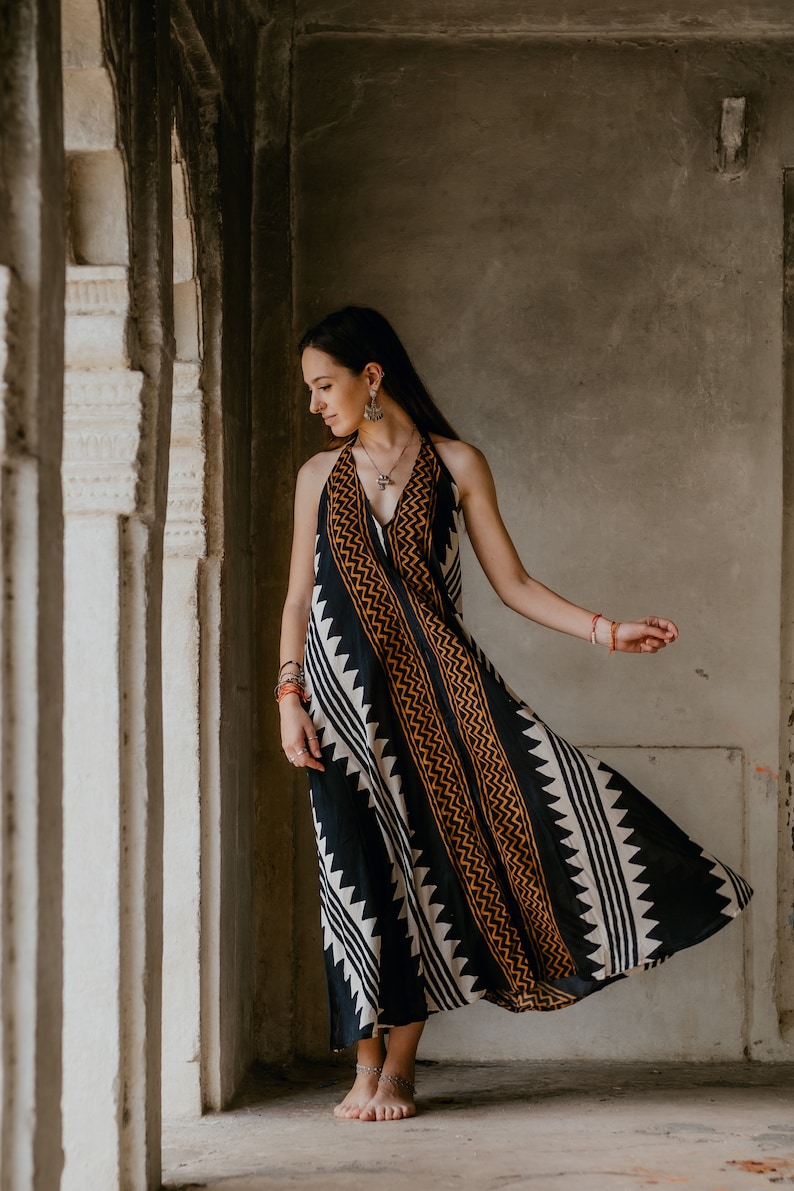May include: A woman wearing a black and white patterned maxi dress with orange accents. The dress has a halter neckline and a flowing skirt. She is standing in a doorway with a concrete wall behind her.
