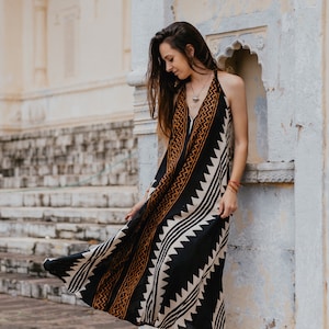May include: A woman wearing a black and white maxi dress with a geometric pattern and brown accents. The dress is sleeveless and has a halter neckline. She is standing in front of a white wall with a decorative archway.