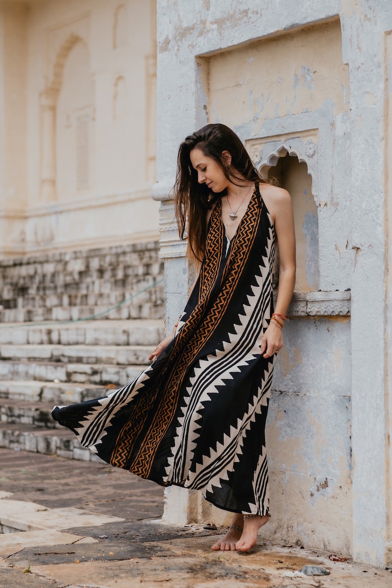 May include: A woman wearing a black and white maxi dress with a geometric pattern and brown accents. The dress is sleeveless and has a halter neckline. She is standing in front of a white wall with a decorative archway.