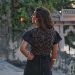 Peut inclure: Une femme portant une robe noire avec une superposition de dentelle et un motif brun. Elle se tient devant une arche en pierre avec une vue sur une &eacute;tendue d'eau en arri&egrave;re-plan.