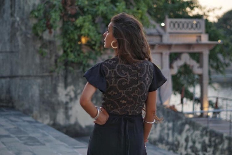 Peut inclure: Une femme portant une robe noire avec une superposition de dentelle et un motif brun. Elle se tient devant une arche en pierre avec une vue sur une &eacute;tendue d'eau en arri&egrave;re-plan.