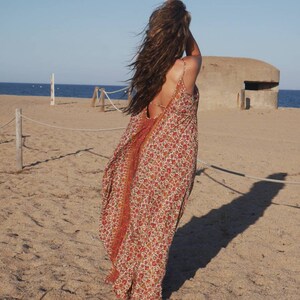 May include: A woman wearing a long, flowing, red and white floral print dress with a low back. The dress is made of a lightweight fabric and has a relaxed fit. The woman is standing on a sandy beach with a concrete bunker in the background.