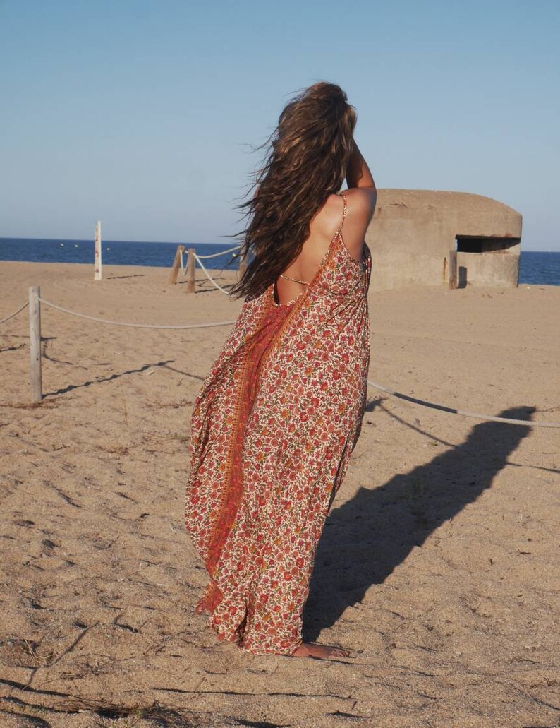 May include: A woman wearing a long, flowing, red and white floral print dress with a low back. The dress is made of a lightweight fabric and has a relaxed fit. The woman is standing on a sandy beach with a concrete bunker in the background.