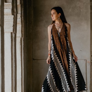 May include: A woman wearing a black and white patterned maxi dress with orange accents. She is standing in a doorway with a concrete wall behind her.