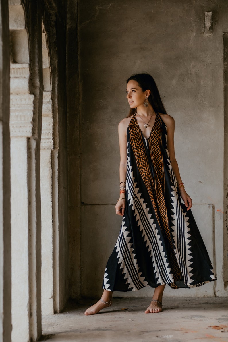 May include: A woman wearing a black and white patterned maxi dress with orange accents. She is standing in a doorway with a concrete wall behind her.