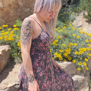 May include: A woman wearing a black and pink floral maxi dress with a lion tattoo on her upper arm. She is standing in front of a rock wall and yellow flowers.
