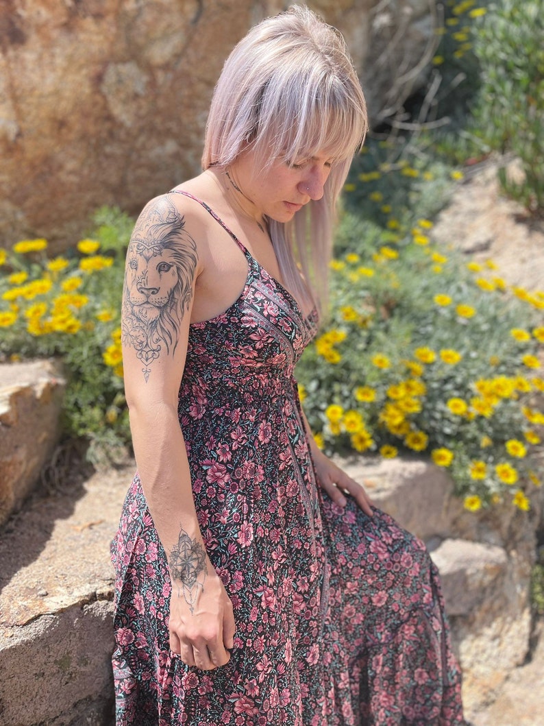May include: A woman wearing a black and pink floral maxi dress with a lion tattoo on her upper arm. She is standing in front of a rock wall and yellow flowers.