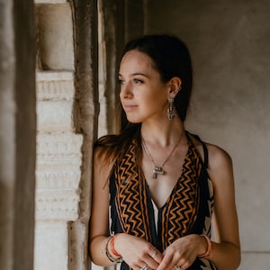 May include: A woman wearing a black and orange patterned dress with a silver necklace and earrings. She is standing in a doorway with a white marble wall behind her.