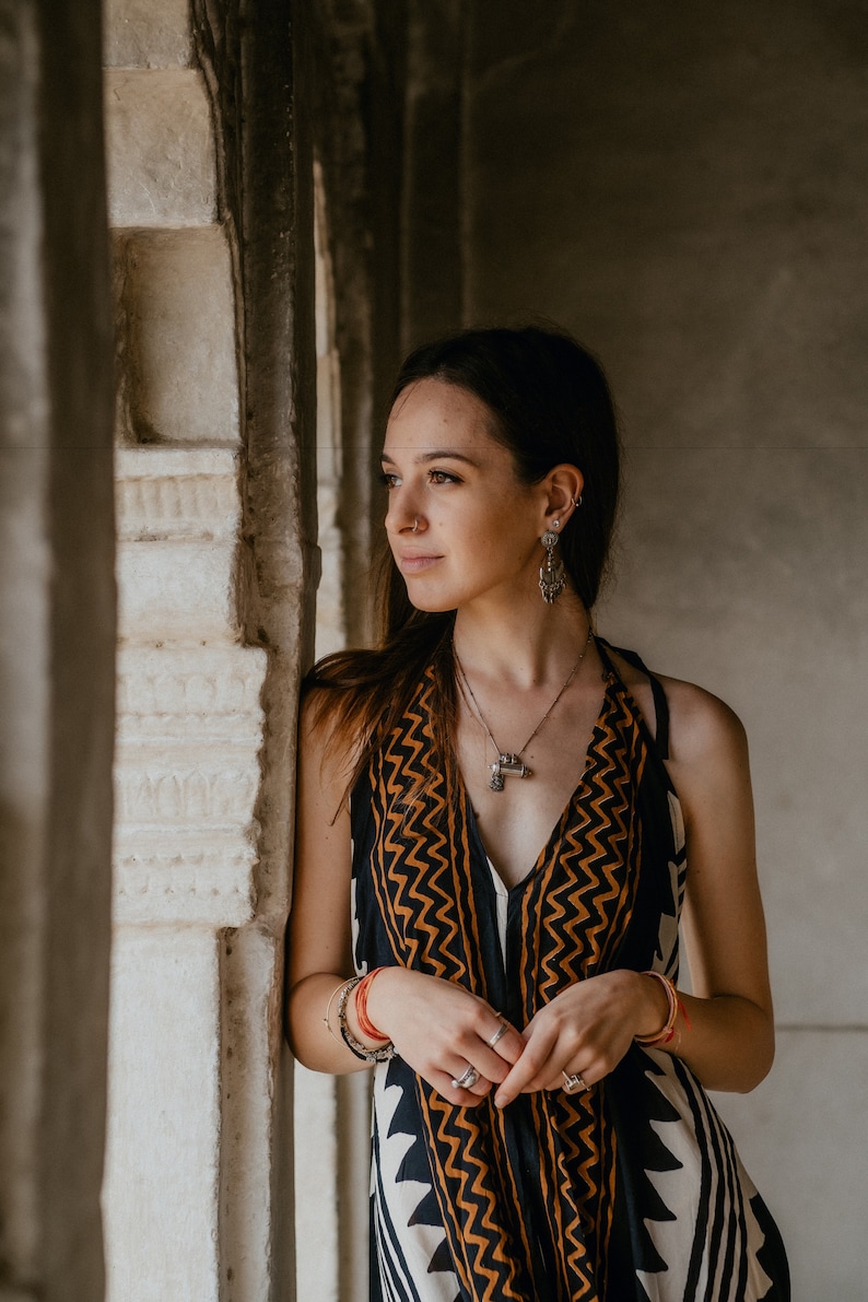 May include: A woman wearing a black and orange patterned dress with a silver necklace and earrings. She is standing in a doorway with a white marble wall behind her.