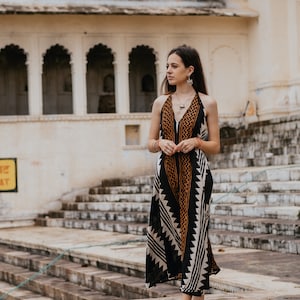 May include: A woman wearing a black and white patterned maxi dress with orange accents stands on a set of stone steps in front of a building with arched doorways.