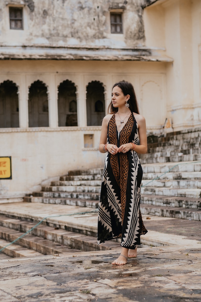 May include: A woman wearing a black and white patterned maxi dress with orange accents stands on a set of stone steps in front of a building with arched doorways.