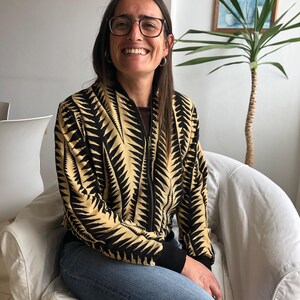 Puede incluir: Una mujer con una chaqueta bomber de patr&oacute;n negro y dorado con una cremallera negra y pantalones vaqueros azules. Est&aacute; sentada en una silla blanca y sonriendo.