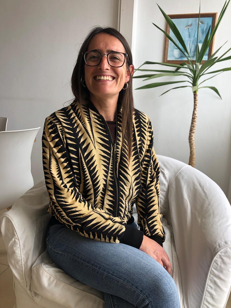 Puede incluir: Una mujer con una chaqueta bomber de patr&oacute;n negro y dorado con una cremallera negra y pantalones vaqueros azules. Est&aacute; sentada en una silla blanca y sonriendo.