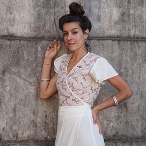 Peut inclure: Une femme portant une robe blanche &agrave; enrouler avec des broderies rouges. Elle se tient devant un mur en b&eacute;ton gris.