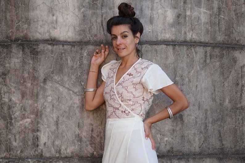 Peut inclure: Une femme portant une robe blanche &agrave; enrouler avec des broderies rouges. Elle se tient devant un mur en b&eacute;ton gris.