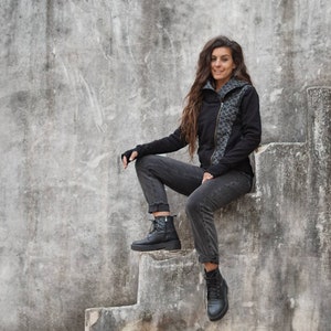 K&ouml;nnte beinhalten: Eine Frau in einer schwarzen Kapuzenjacke mit Rei&szlig;verschluss und einem Muster sitzt auf einer Betontreppe. Sie tr&auml;gt eine schwarze Jeans und schwarze Stiefel.