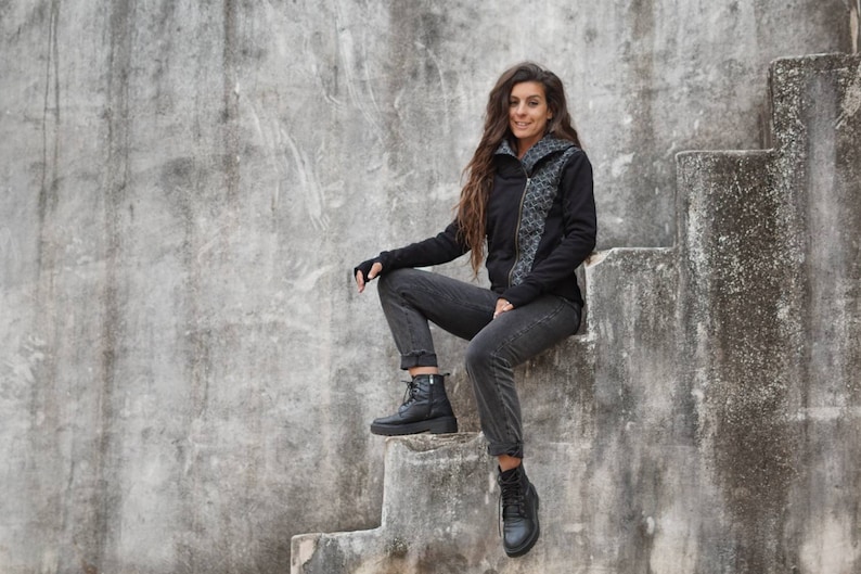 K&ouml;nnte beinhalten: Eine Frau in einer schwarzen Kapuzenjacke mit Rei&szlig;verschluss und einem Muster sitzt auf einer Betontreppe. Sie tr&auml;gt eine schwarze Jeans und schwarze Stiefel.