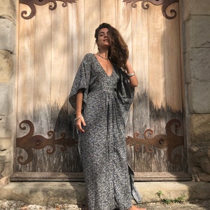 May include: A woman wearing a long, black and white floral print maxi dress with a V-neckline and flowing sleeves. She is standing in front of a wooden door with iron hinges.
