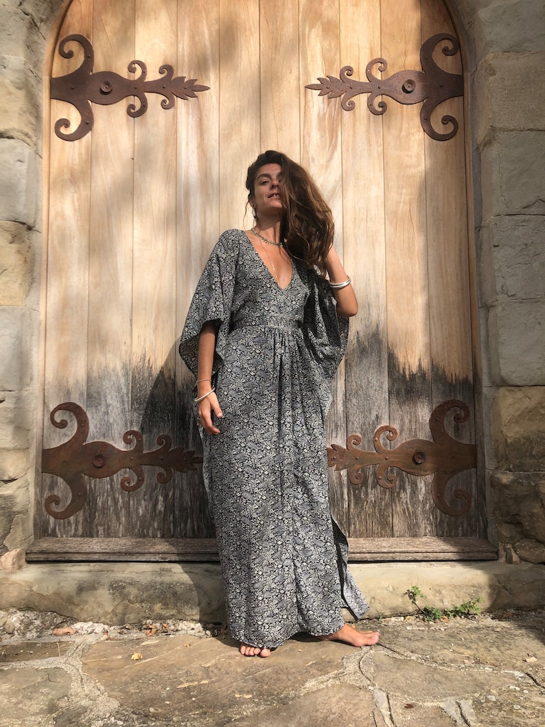 May include: A woman wearing a long, black and white floral print maxi dress with a V-neckline and flowing sleeves. She is standing in front of a wooden door with iron hinges.