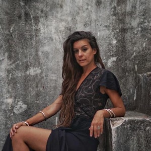Peut inclure: Une femme aux longs cheveux bruns portant une robe en dentelle noire et des bracelets argent&eacute;s est assise sur un escalier en b&eacute;ton.