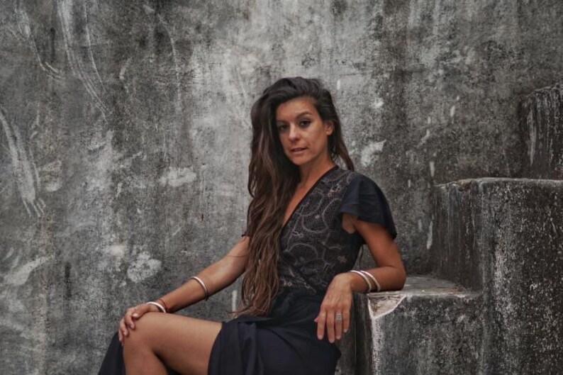 Peut inclure: Une femme aux longs cheveux bruns portant une robe en dentelle noire et des bracelets argent&eacute;s est assise sur un escalier en b&eacute;ton.