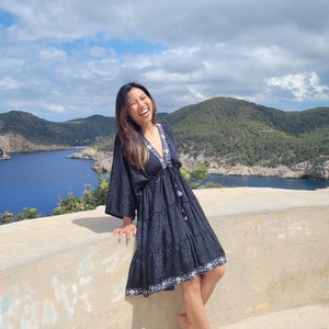 May include: A woman wearing a black tiered dress with white embroidery and a blue trim, standing on a concrete platform overlooking a body of water.