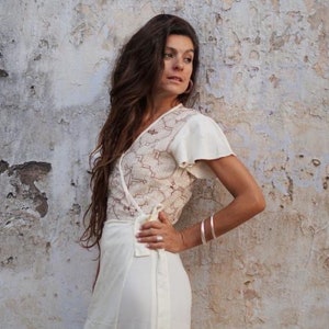 Peut inclure: Une femme portant une robe blanche enroul&eacute;e avec un motif de dentelle d&eacute;licat. La robe a des manches courtes et un col en V. Elle se tient devant un mur blanc textur&eacute;.