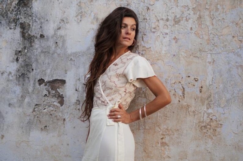 Peut inclure: Une femme portant une robe blanche enroul&eacute;e avec un motif de dentelle d&eacute;licat. La robe a des manches courtes et un col en V. Elle se tient devant un mur blanc textur&eacute;.