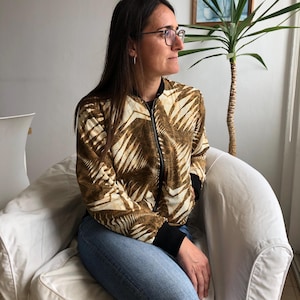Puede incluir: Una mujer con una chaqueta bomber de color marr&oacute;n y blanco con un patr&oacute;n geom&eacute;trico. Est&aacute; sentada en un sill&oacute;n blanco y mirando hacia la derecha.