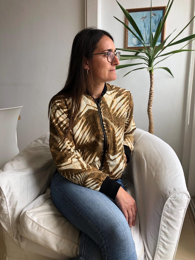 Puede incluir: Una mujer con una chaqueta bomber de color marr&oacute;n y blanco con un patr&oacute;n geom&eacute;trico. Est&aacute; sentada en un sill&oacute;n blanco y mirando hacia la derecha.