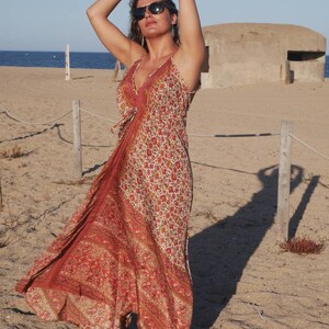 May include: A woman wearing a long, red and white floral print dress with a halter neckline. She is standing on a sandy beach with a concrete bunker in the background.